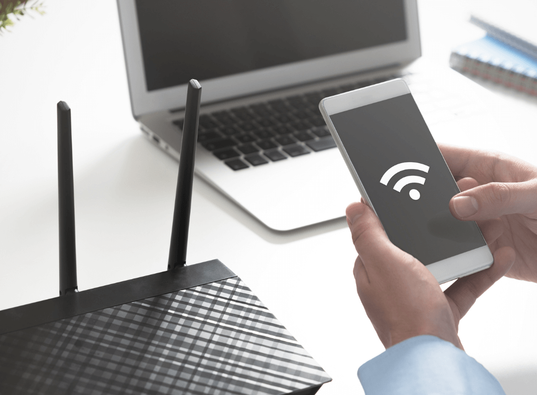 Smartphone with Wi-Fi symbol held beside laptop and router.